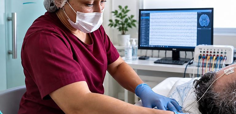 Julia Alcantara realizando montagem de eletrodos para EEG, com monitor exibindo o traçado do exame